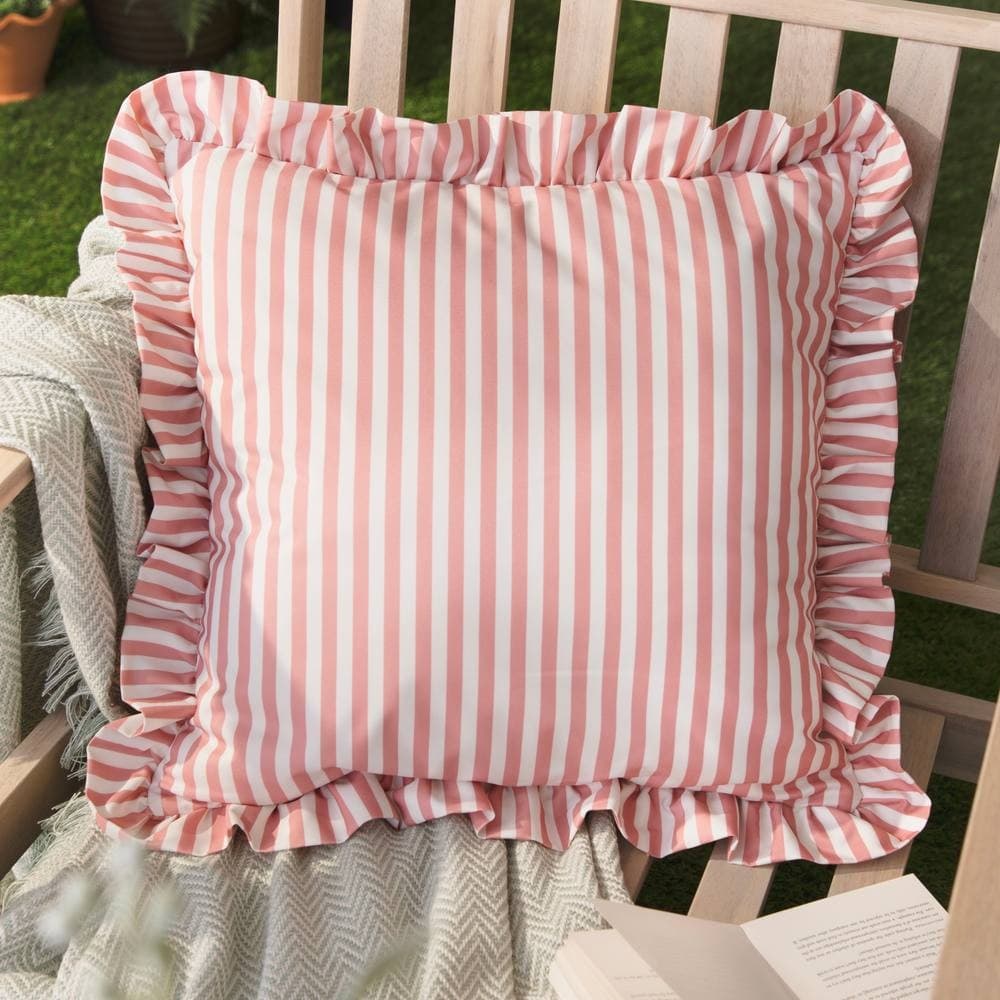 Pink and white striped outdoor cushion with a ruffled edge, made of durable fabric, placed on a wooden bench with a cozy throw and book nearby.