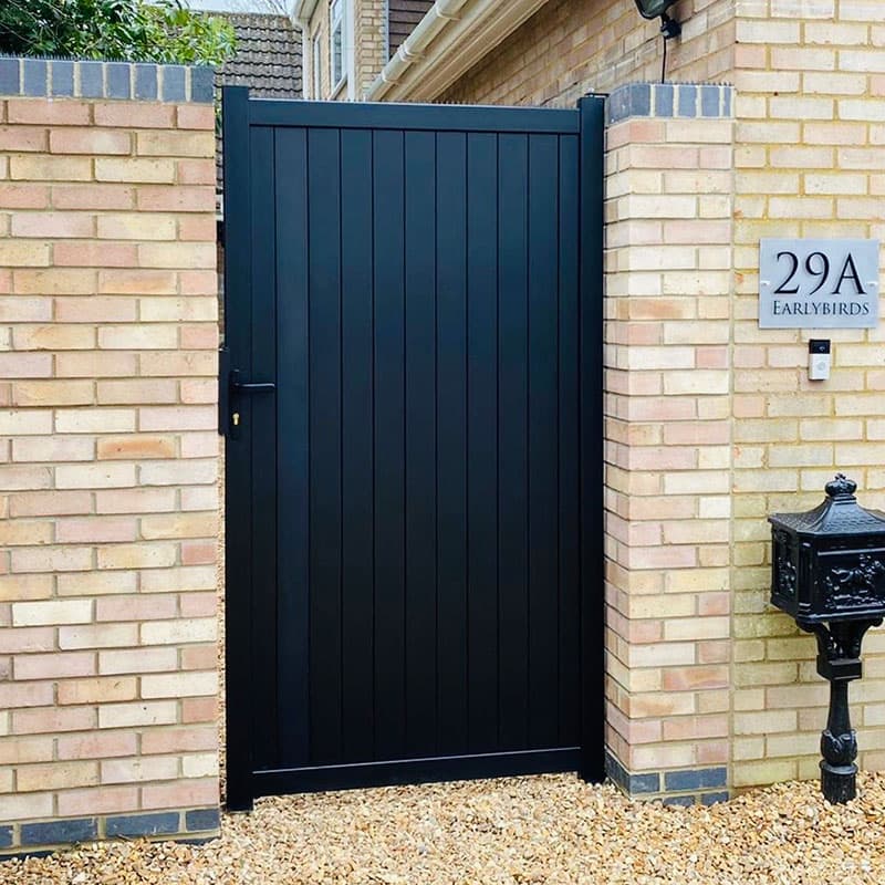 Black premium metal side gate with vertical slats, featuring a sleek design and sturdy latch, set against a brick wall and gravel pathway.