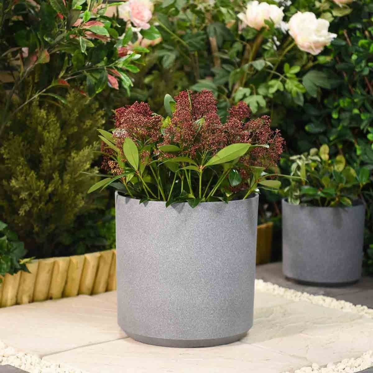 Grey 31cm planter made of textured material, featuring a rounded design and filled with lush green plants and burgundy flowers, set against a lush garden backdrop.