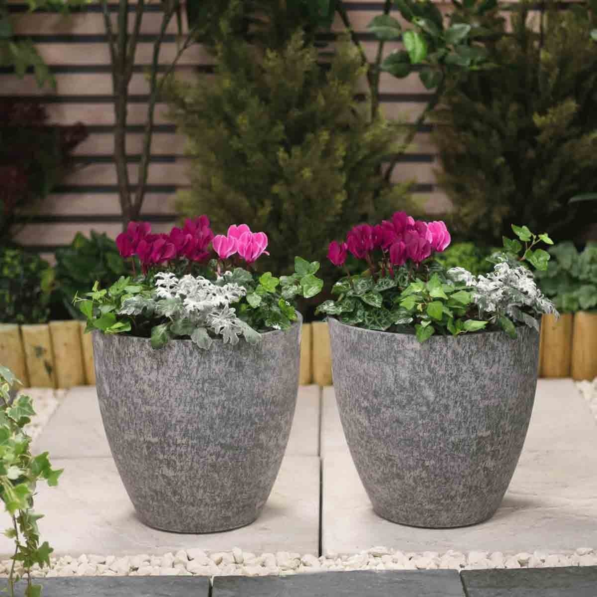 Two stone effect grey planters, 28 cm in size, filled with vibrant pink and white flowers, set against a lush garden backdrop.