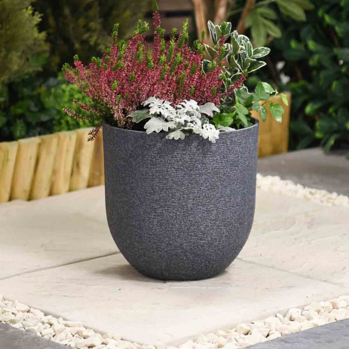 Stone effect grey planter, 26cm in diameter, featuring a textured surface, filled with vibrant green and purple plants, set on a stone patio surrounded by greenery.