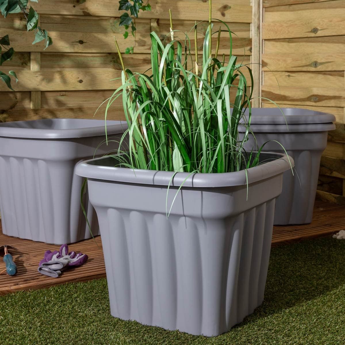 Grey 49cm square planter made from durable plastic, featuring ribbed sides and filled with lush green grass, set against a wooden backdrop.