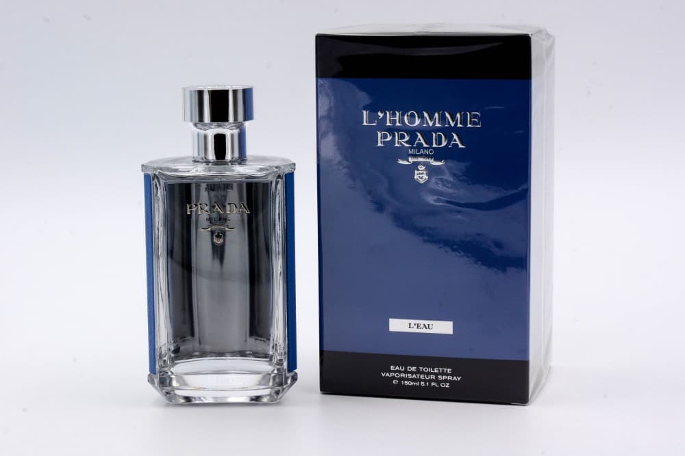 Clear glass bottle with a silver cap, featuring a sleek design and engraved "Prada" logo, alongside a blue box labeled "L'Homme Prada Milano L'Eau."