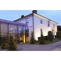 White two-story spa building with glass extension, surrounded by greenery, at dusk.