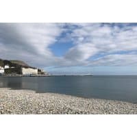 Pebbled beach with calm sea, distant hotel, and cloudy sky in North Wales for seaside stay package with Prosecco.