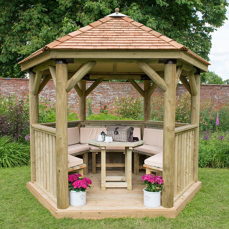 Luxury wooden gazebo with a New England cedar roof, featuring light-colored wooden beams, cushioned seating for up to 10, and flower pots at the base, set in a lush garden.