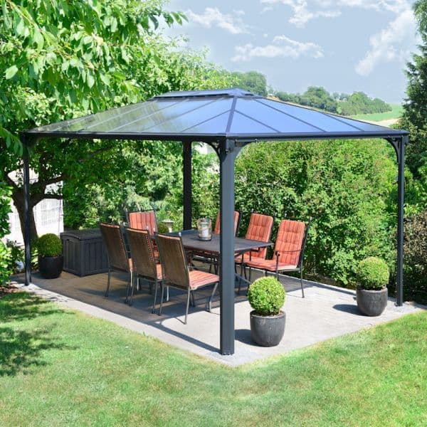 A spacious 14'x10' Palram Canopia Martinique gazebo with a dark metal frame, clear polycarbonate roof, and a patio area featuring a dining set with red cushioned chairs and potted plants.