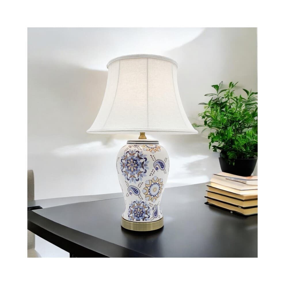 Elegant white table lamp with a floral blue and gold ceramic base, featuring a white fabric shade, measuring L20 x W20 x H45 cm, set on a dark table beside stacked books and a plant.