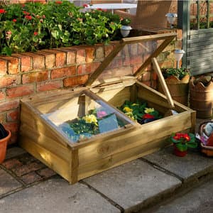Large wooden cold frame in natural wood finish, featuring a sloped glass lid, housing vibrant flowers, set against a brick wall with potted plants and greenery.