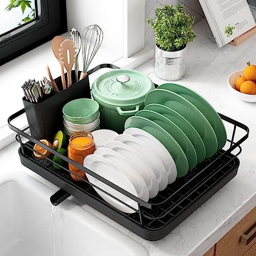 Space-saving black stainless steel dish drainer with white and green plates, a cutlery holder, and kitchen utensils, set on a marble countertop next to a window with plants.
