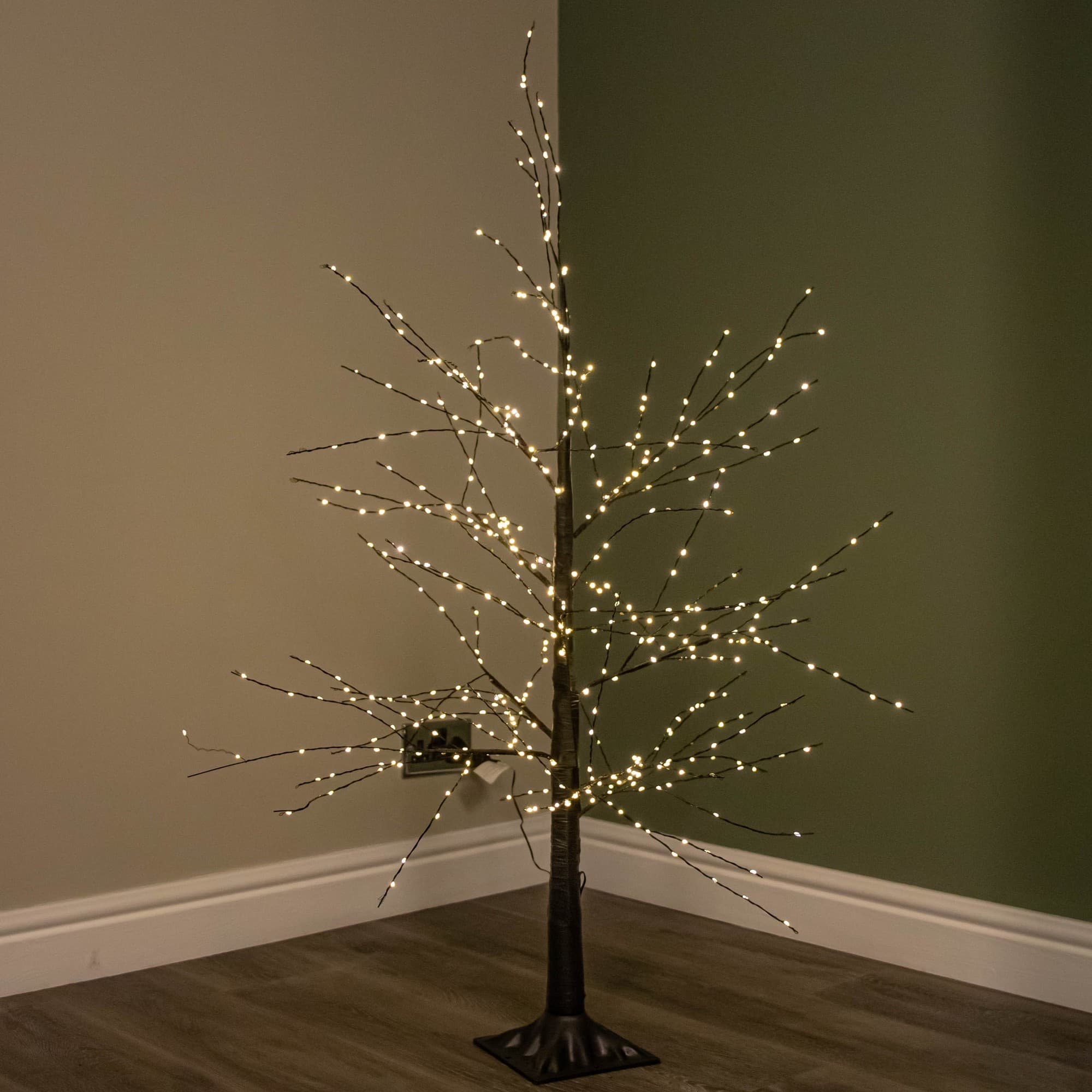4ft black micro dot Christmas tree with a slender trunk and bare branches, adorned with 500 warm white LEDs that twinkle and shine, set against a muted green wall.