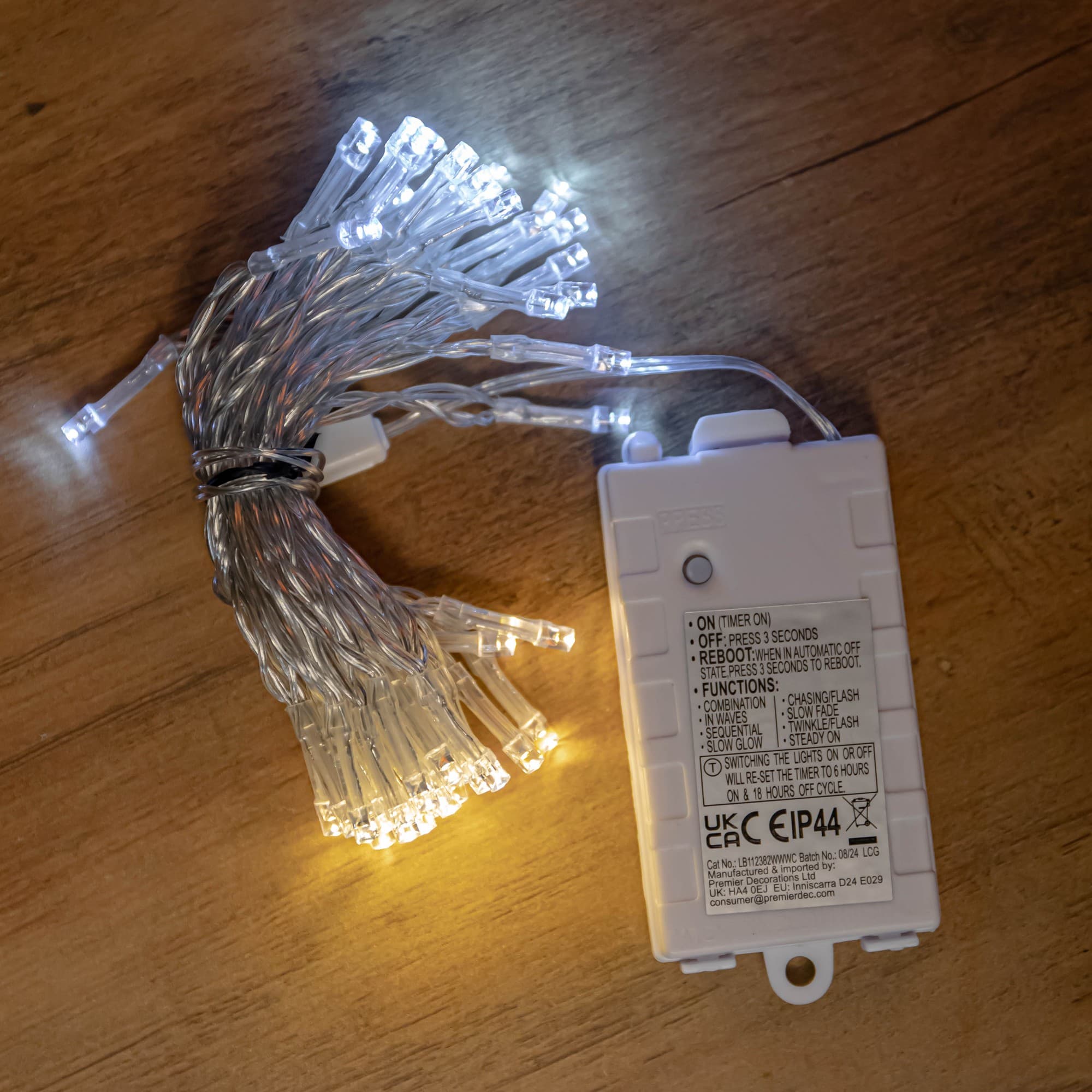 Battery-operated Christmas string lights featuring 50 LED bulbs in cool and warm white, with a clear plastic casing and a timer function, resting on a wooden surface.