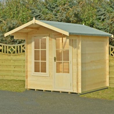 A wooden log cabin summerhouse with a light wood finish, featuring a sloped grey roof, double front doors, and large windows, set against a green fence and trees.