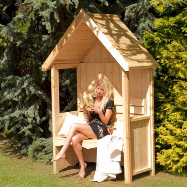 Natural wood garden arbour seat with a slanted roof, featuring a spacious bench and a woman reading, surrounded by lush greenery.