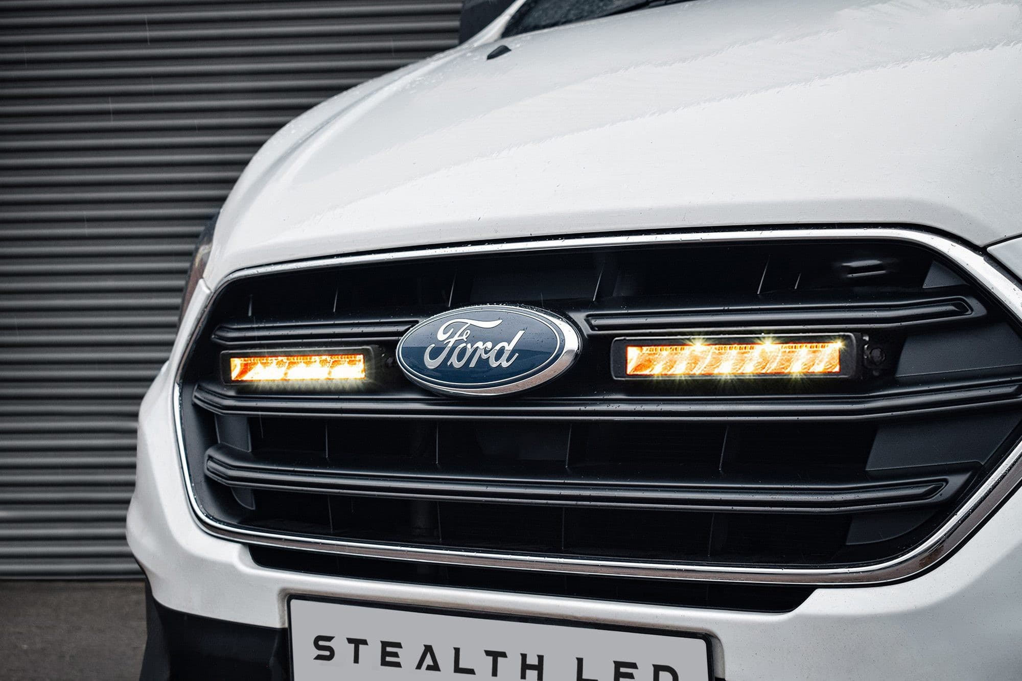 White Ford Transit Custom grille featuring a sleek black design with twin 10" flashing luminous LED lights, showcasing bright orange illumination and a prominent Ford logo.