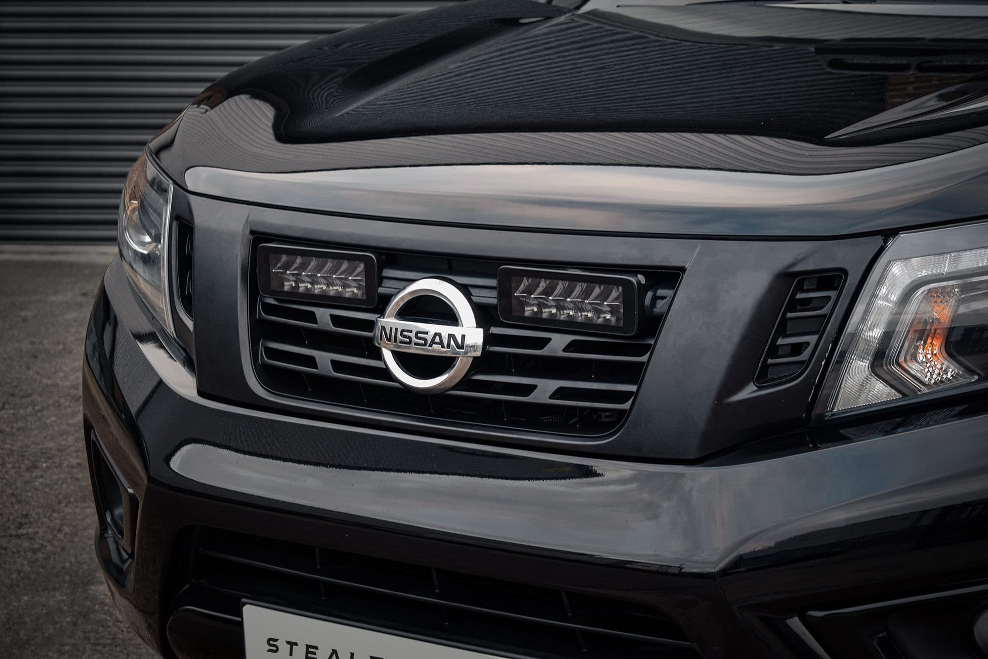 Black Nissan Navara D23 front grille featuring a chrome Nissan emblem, integrated with twin 7" luminous LED lights, showcasing a sleek design and high brightness of 10,080 lumens.