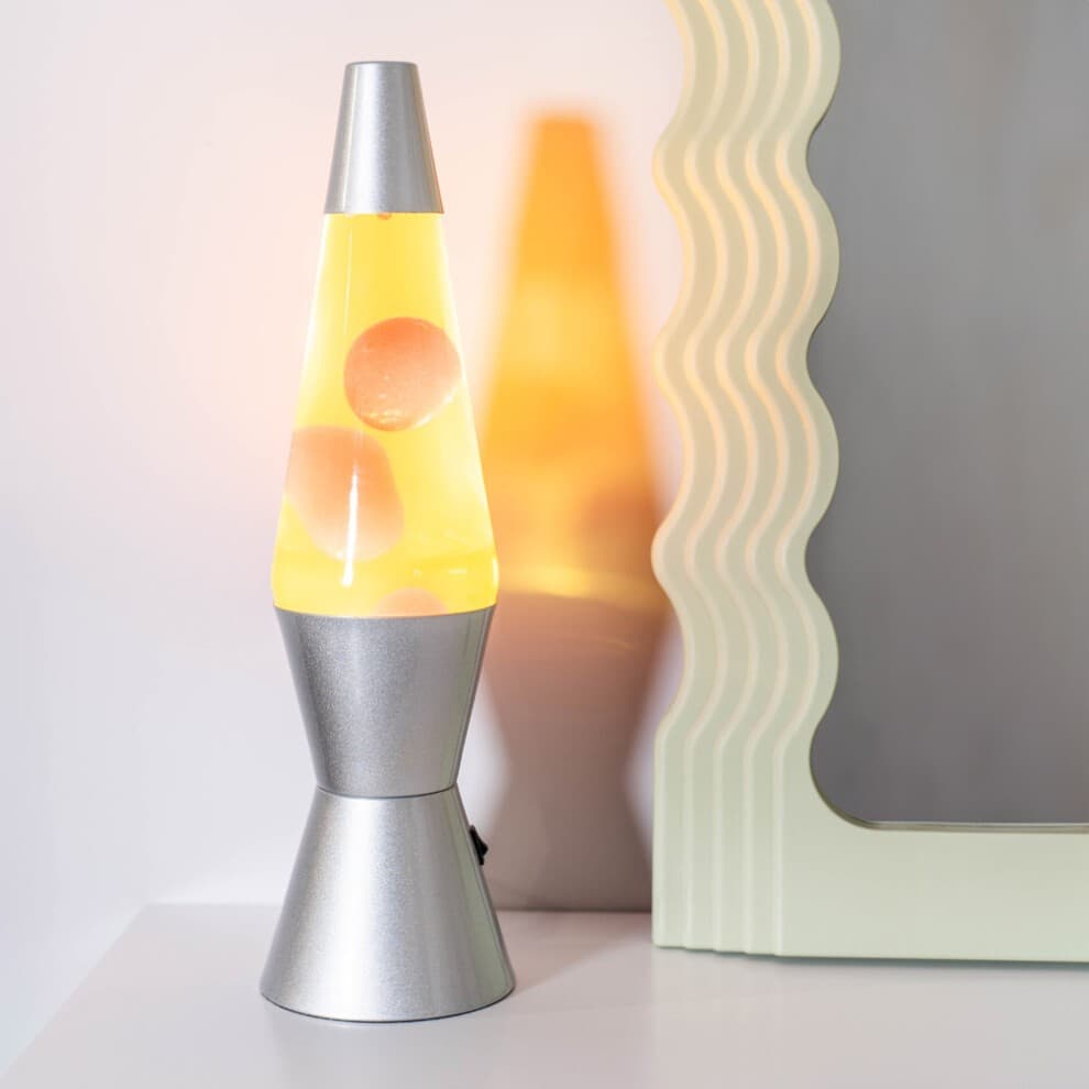 Retro lava lamp featuring a silver base, with yellow liquid and red lava bubbles, casting a warm glow beside a wavy-edged mirror.