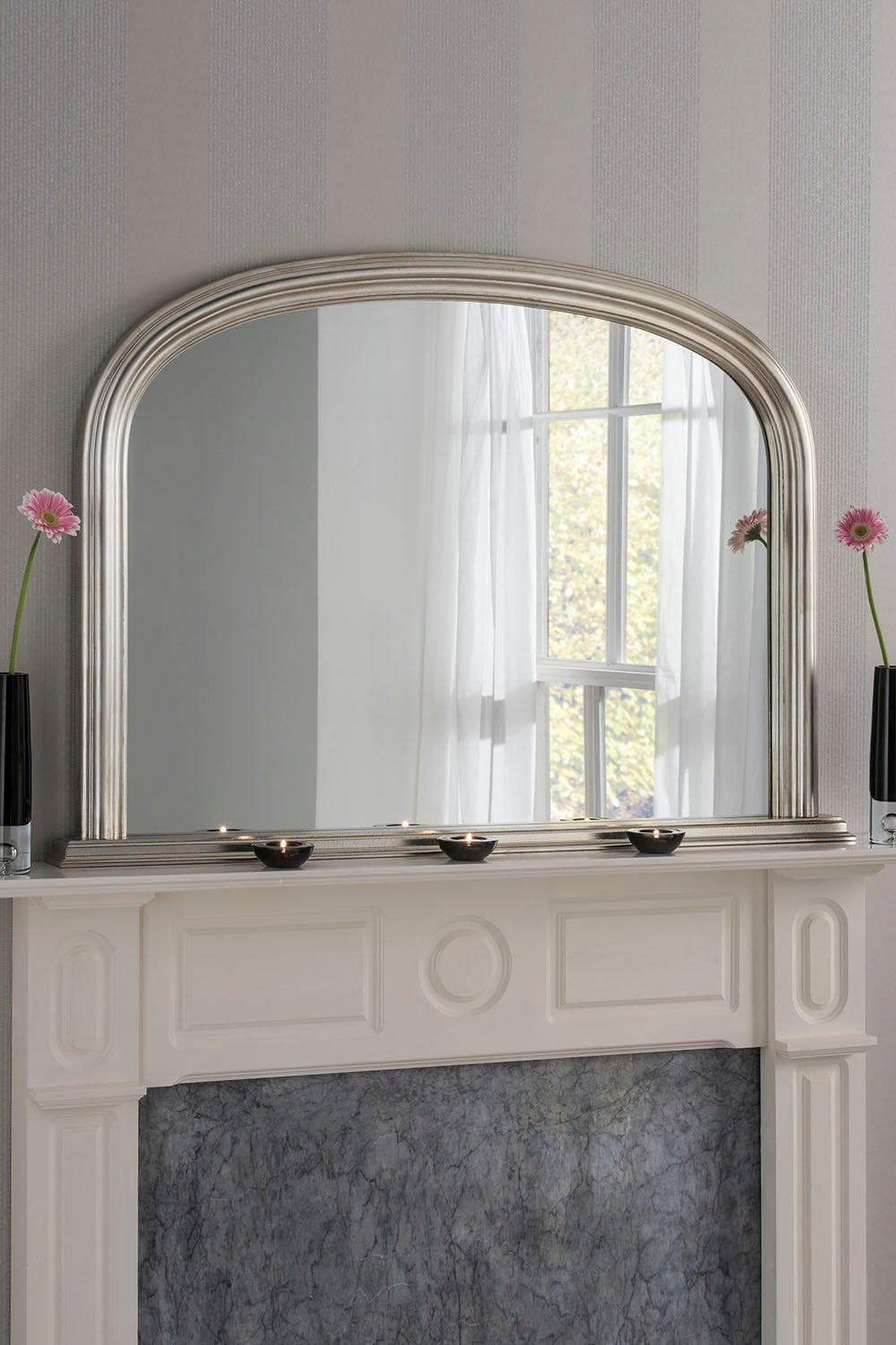 Arched contemporary mirror in metallic silver, measuring 112cm wide by 77cm high, featuring a sleek frame above a marble mantel with decorative vases and flowers.