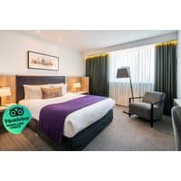 Hotel room with a double bed, purple throw, two bedside lamps, armchair, and large window with dark curtains.
