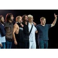 Band members standing together on stage, wearing casual clothing, with one member raising an arm in a gesture.