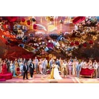"La Traviata opera scene with vibrant costumes, large ensemble cast, and elaborate stage set with red and gold decor"