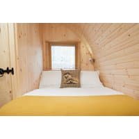 Wooden glamping pod interior with double bed, yellow blanket, and decorative pillow under a small window.
