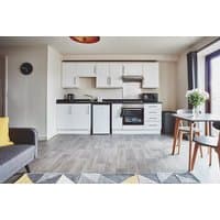 Modern studio apartment with white cabinets, compact kitchen, dining table, and sofa, featuring wood flooring.