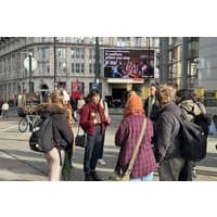 Group of people standing outdoors in a city, with backpacks and jackets, near a large digital billboard.