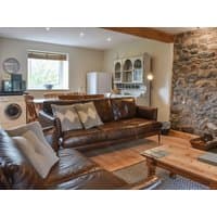 Cozy cottage living room with brown leather sofas, stone wall, wooden coffee table, and kitchen area with appliances.