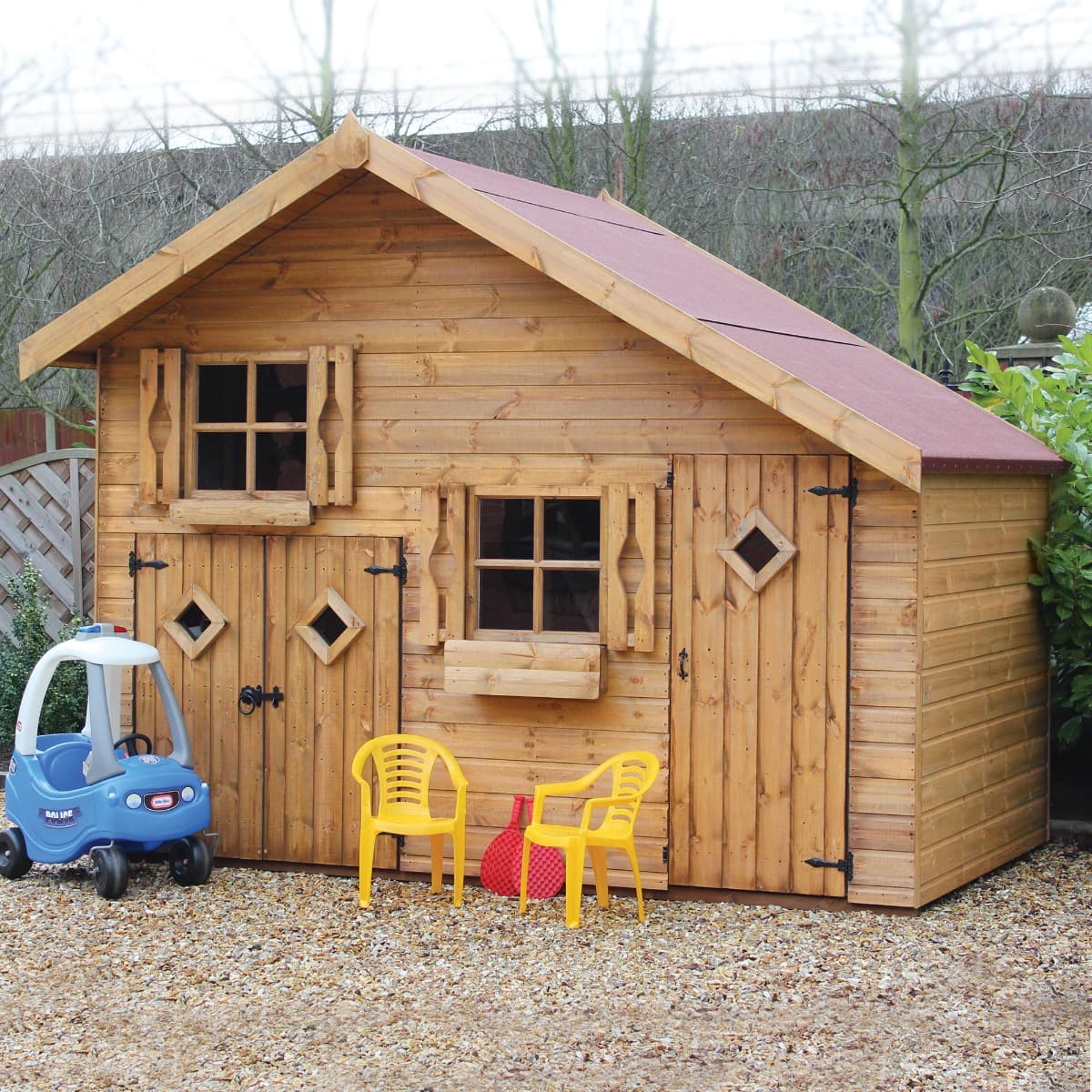 Traditional wooden playhouse in natural wood finish, featuring a red roof, two storeys, garage door, diamond-shaped windows, and yellow chairs outside on gravel.