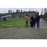 Group of people in winter clothing standing on cobblestone near historic building and large cannon in a park setting.