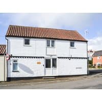 White two-story cottage with red-tiled roof, small front windows, and a central door, located on a street corner.