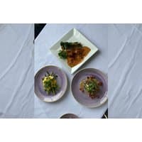 Plated gourmet meals on white and purple dishes, featuring greens and garnishes, on a white tablecloth.