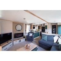 Modern caravan interior with grey sofa, flat-screen TV, electric fireplace, and kitchen area with teal cabinets.