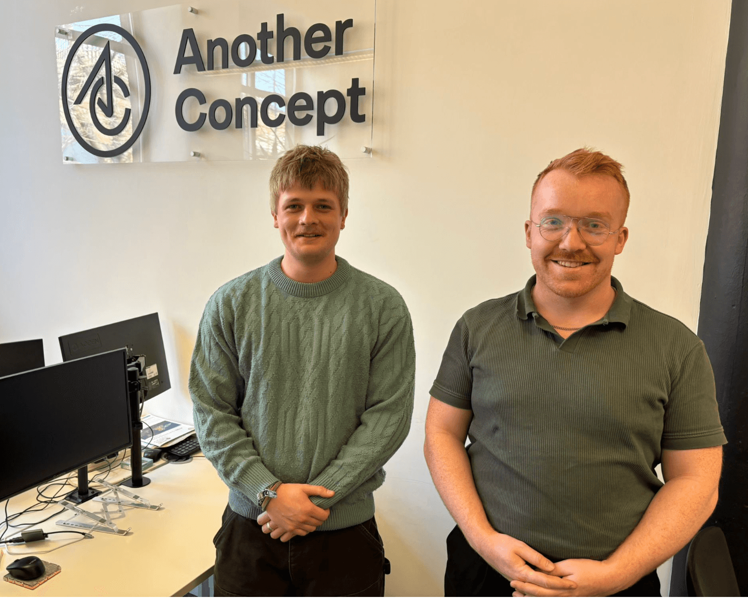 Two young men stand in an office space, smiling beside a sign for "Another Concept," with computers in the background.