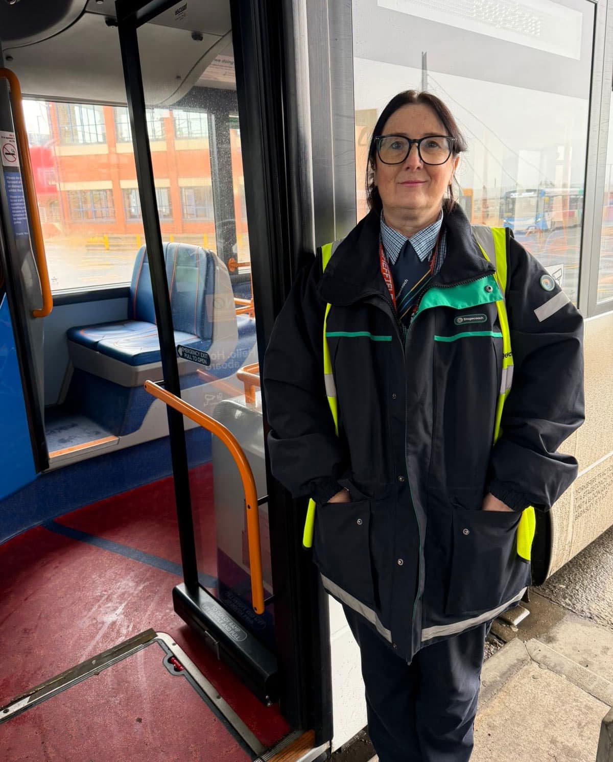 Barnsley bus driver stands proudly by her bus, wearing a uniform and reflective vest, with a city backdrop in a rainy atmosphere.