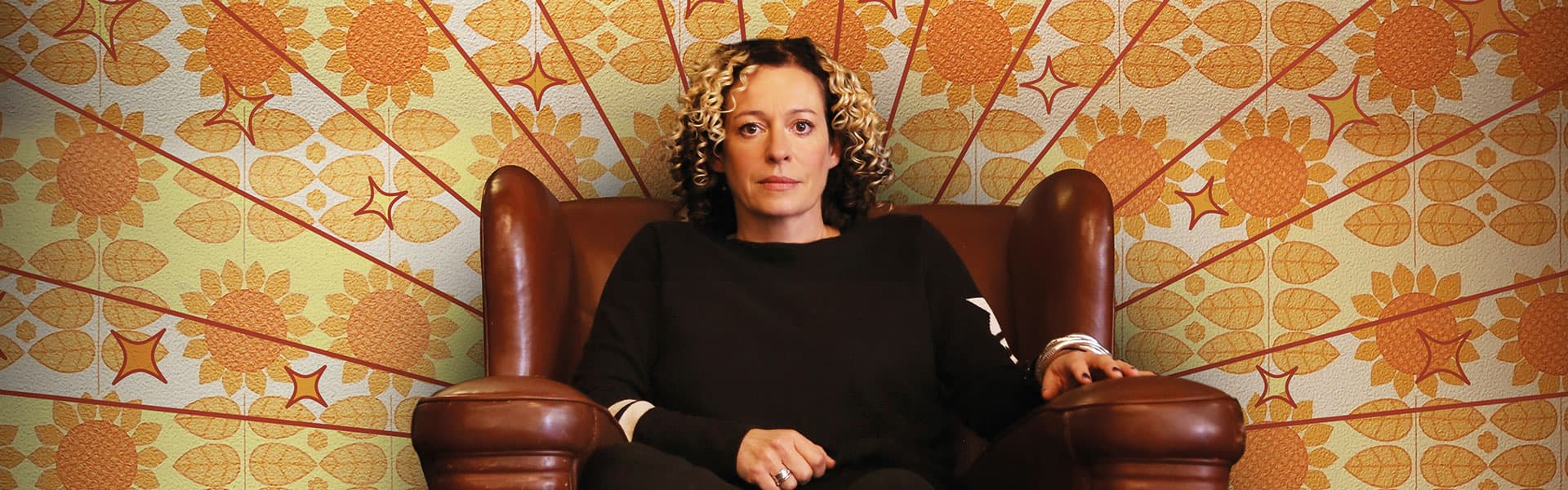 A woman with curly hair sits confidently in a brown leather armchair against a vibrant floral-patterned backdrop.