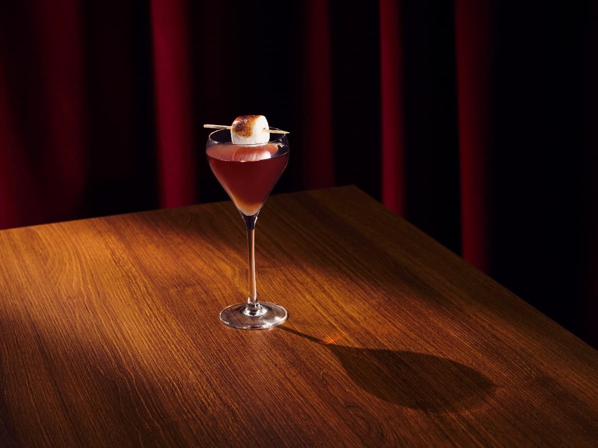A stylish cocktail with a toasted marshmallow garnish sits on a wooden table, illuminated by soft, warm lighting.