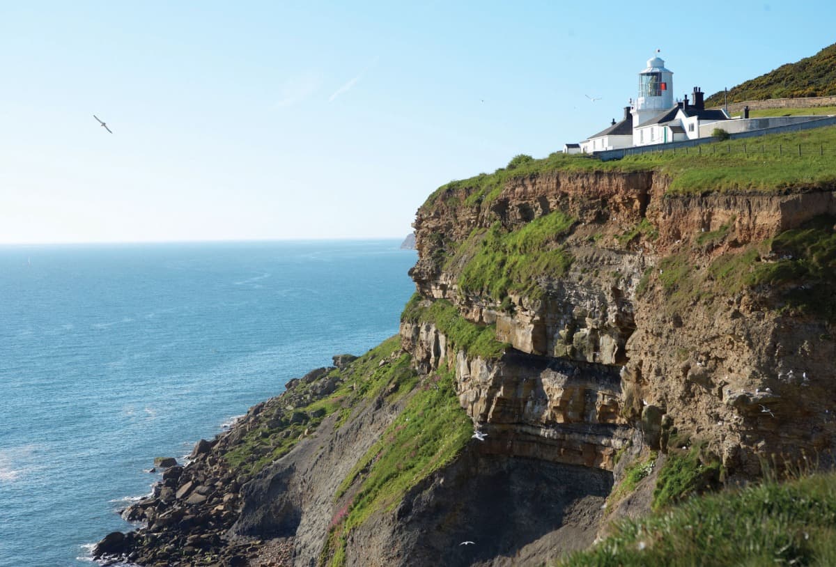 Clifftop lighthouse overlooking the sea, surrounded by lush green hills and a clear blue sky, near the Yorkshire coast.