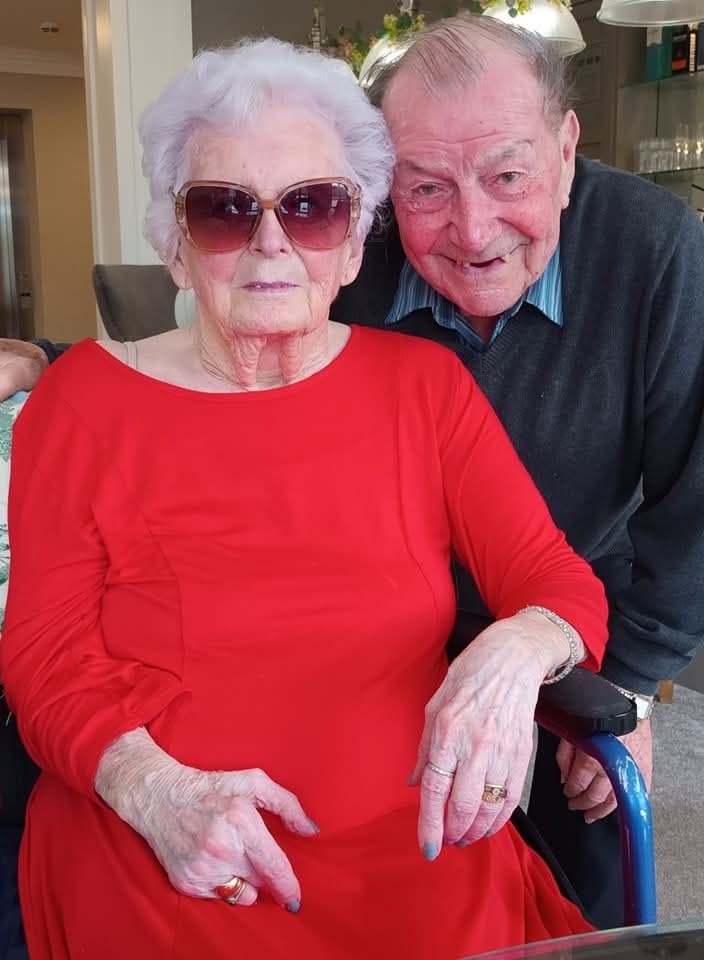 Elderly couple in a cozy setting, the woman in a red dress and sunglasses, smiling with love and warmth.