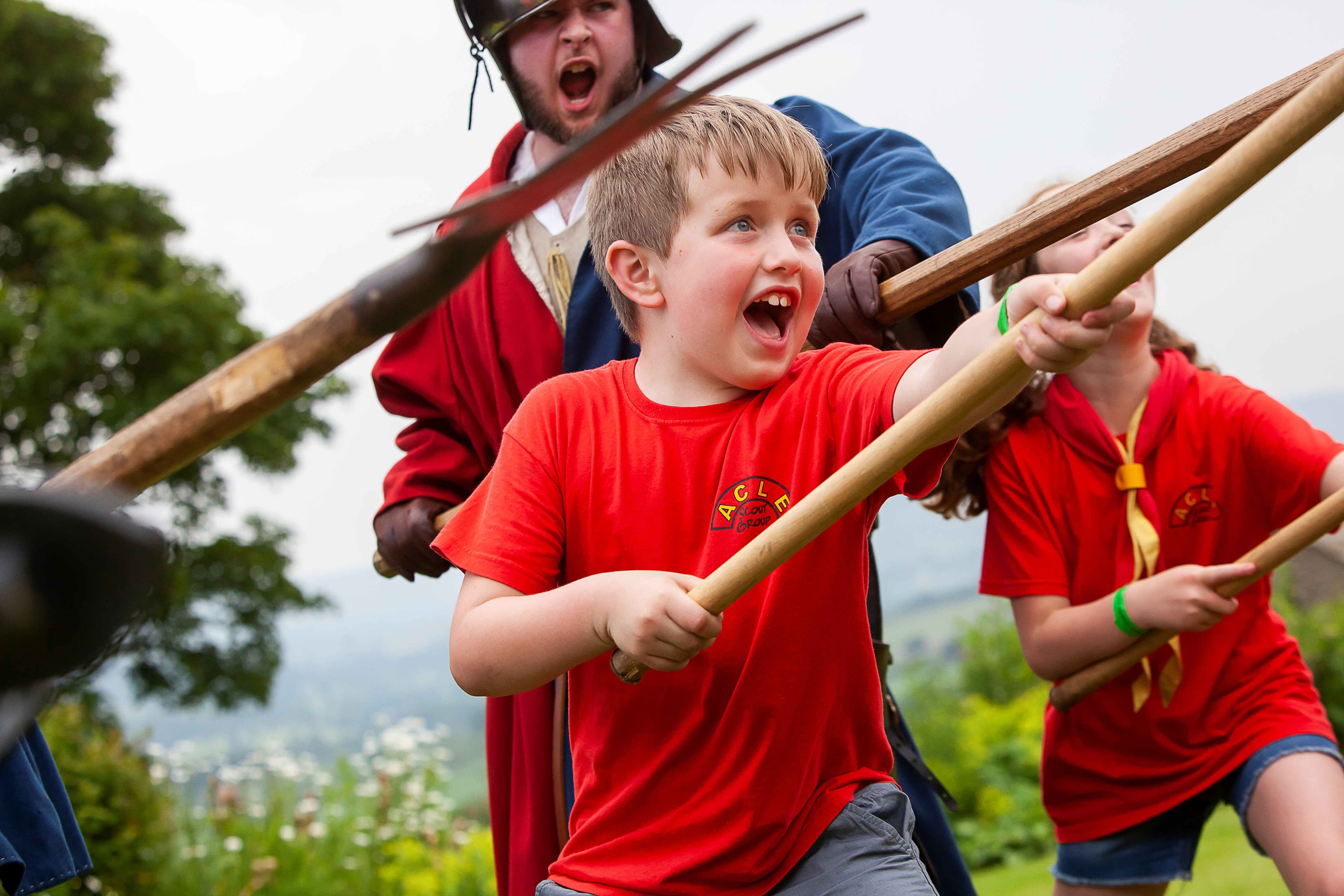 Children in bright red shirts enthusiastically engage in a mock battle at Bolton Castle, surrounded by lush greenery.