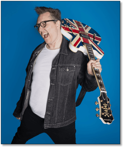 A musician with glasses joyfully poses against a bright blue background, holding a Union Jack guitar.