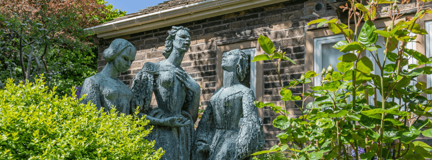 Three bronze statues of the Brontë sisters stand among vibrant greenery outside the Brontë Parsonage Museum in Yorkshire.
