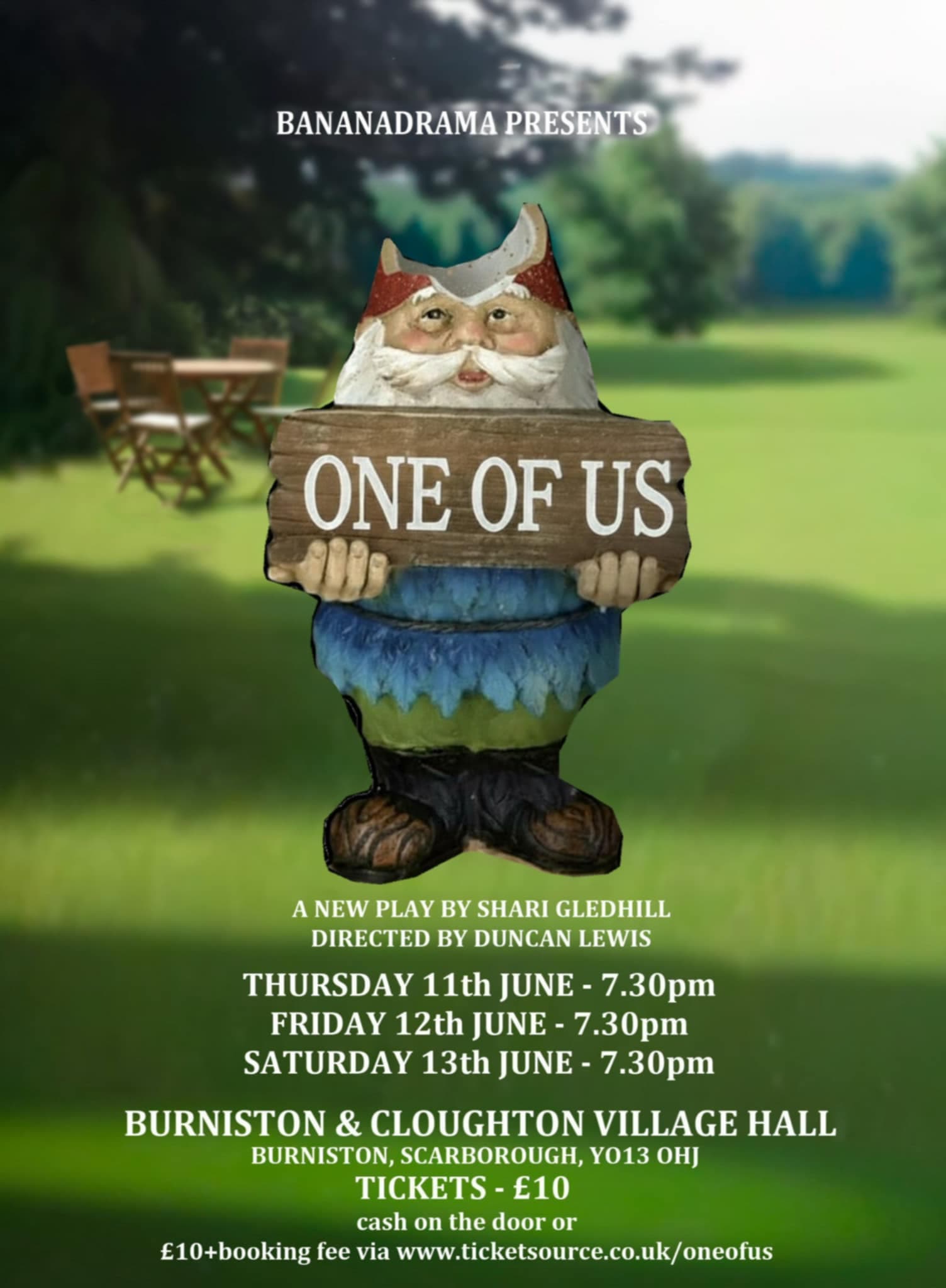 A whimsical gnome statue holds a sign reading "One of Us" outside Burniston and Cloughton Village Hall, set in a green landscape.