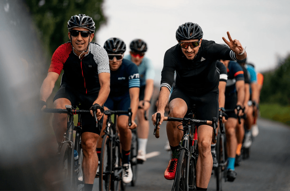 Cyclists riding on a scenic road near Grantley Hall, smiling and enjoying camaraderie under a bright sky.