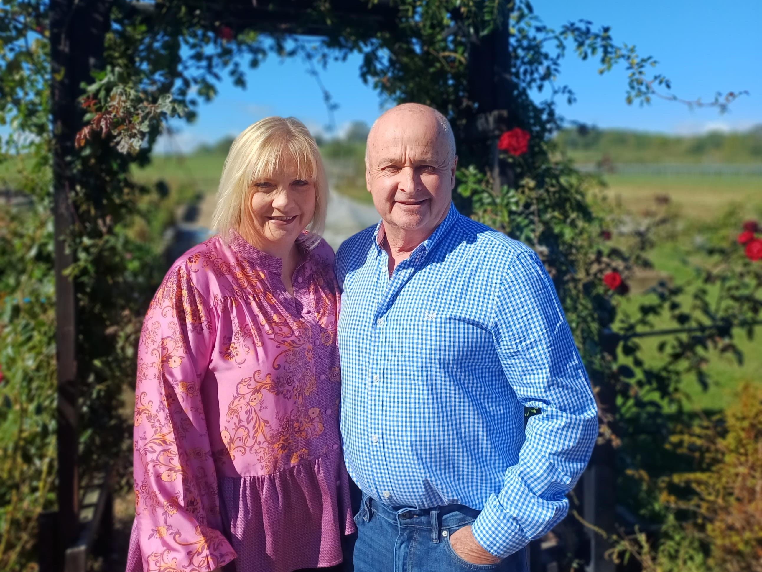 Smiling couple stands in front of a floral archway, with lush greenery and blue skies in the background, near Pickering.