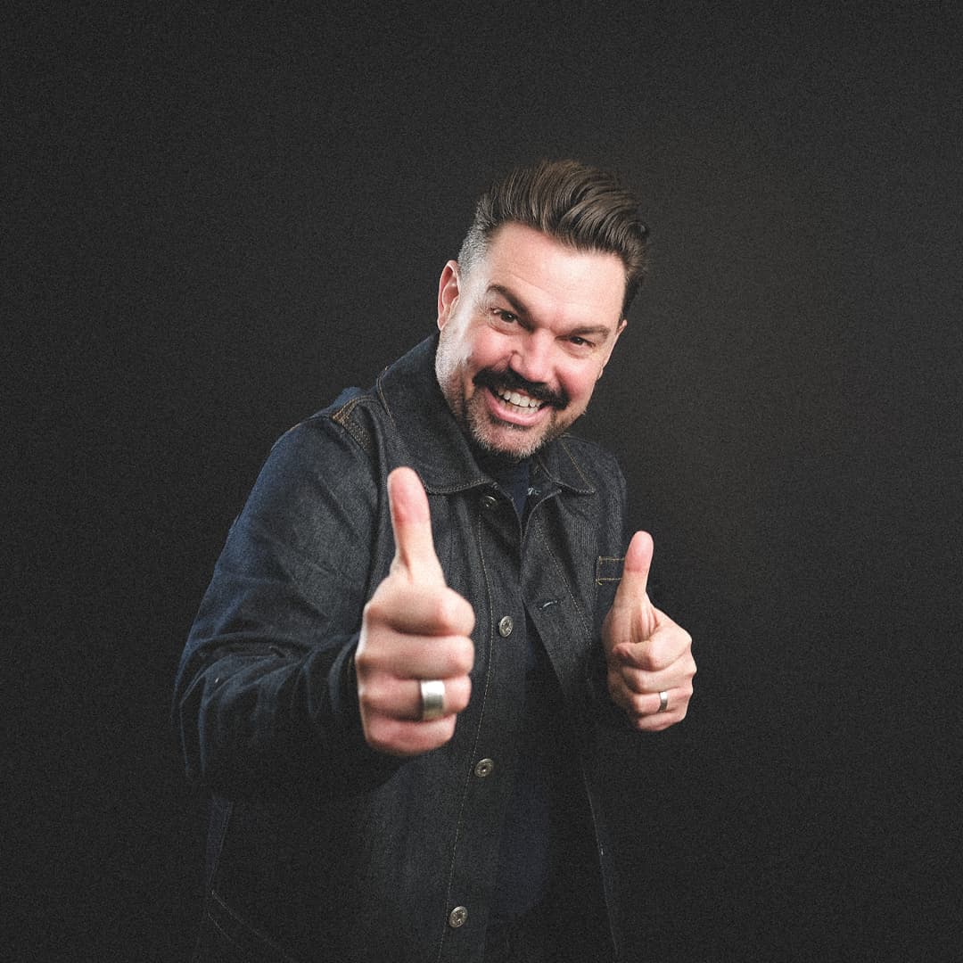 Comedian Charlie Baker smiles and gives a thumbs-up, wearing a denim jacket against a dark backdrop.
