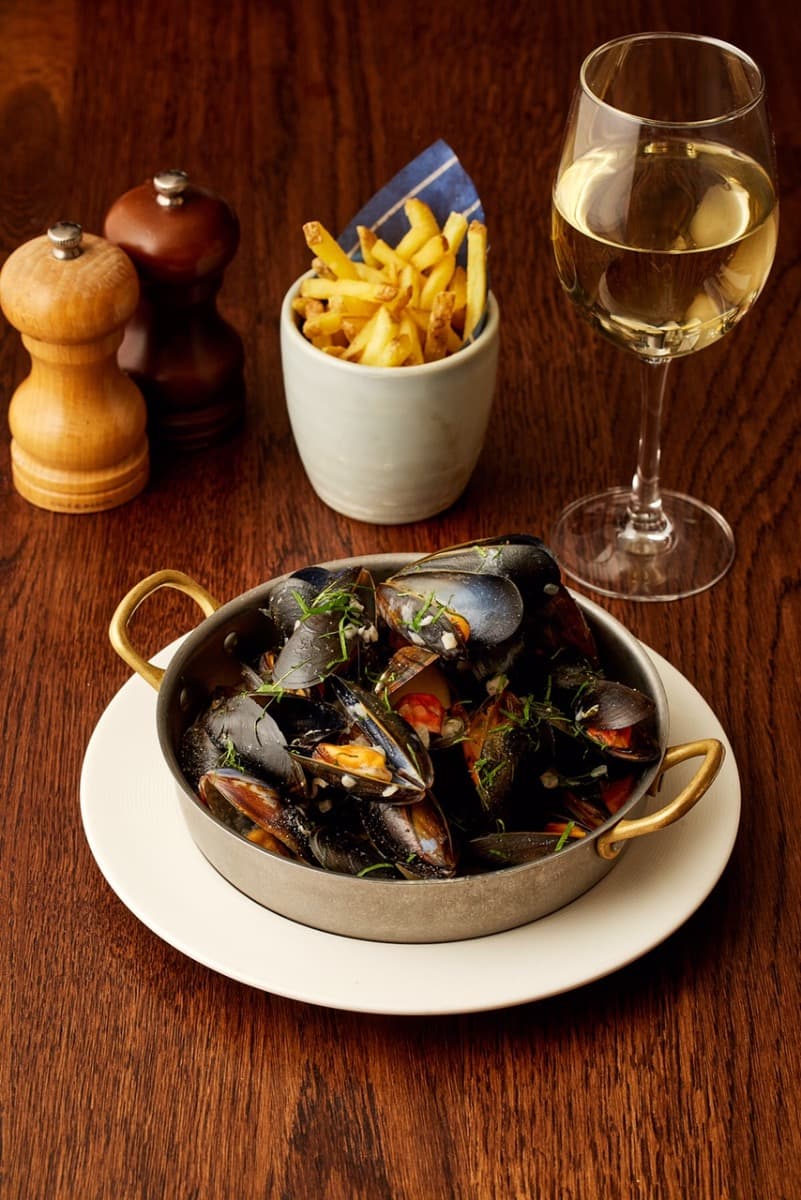 A bowl of mussels in a rich broth, served with fries and a glass of white wine on a wooden table at Côte Brasserie.