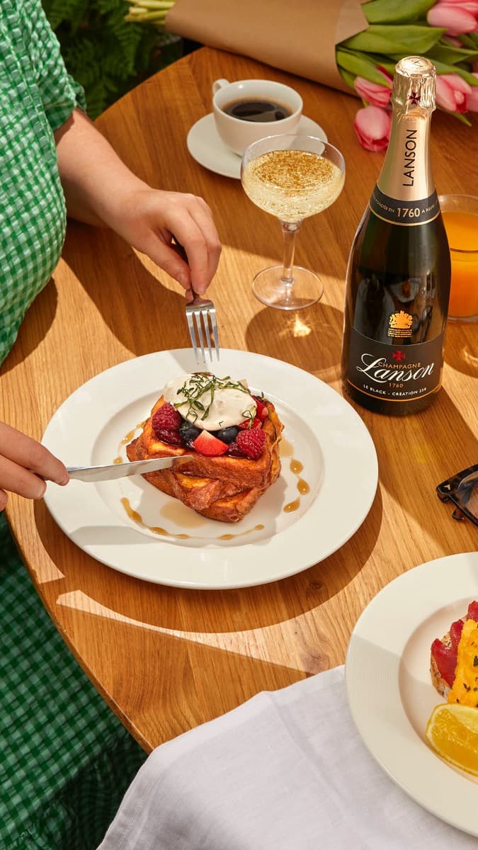 A hand cutting into a decadent dessert topped with berries, beside a glass of champagne and a coffee, set on a wooden table.
