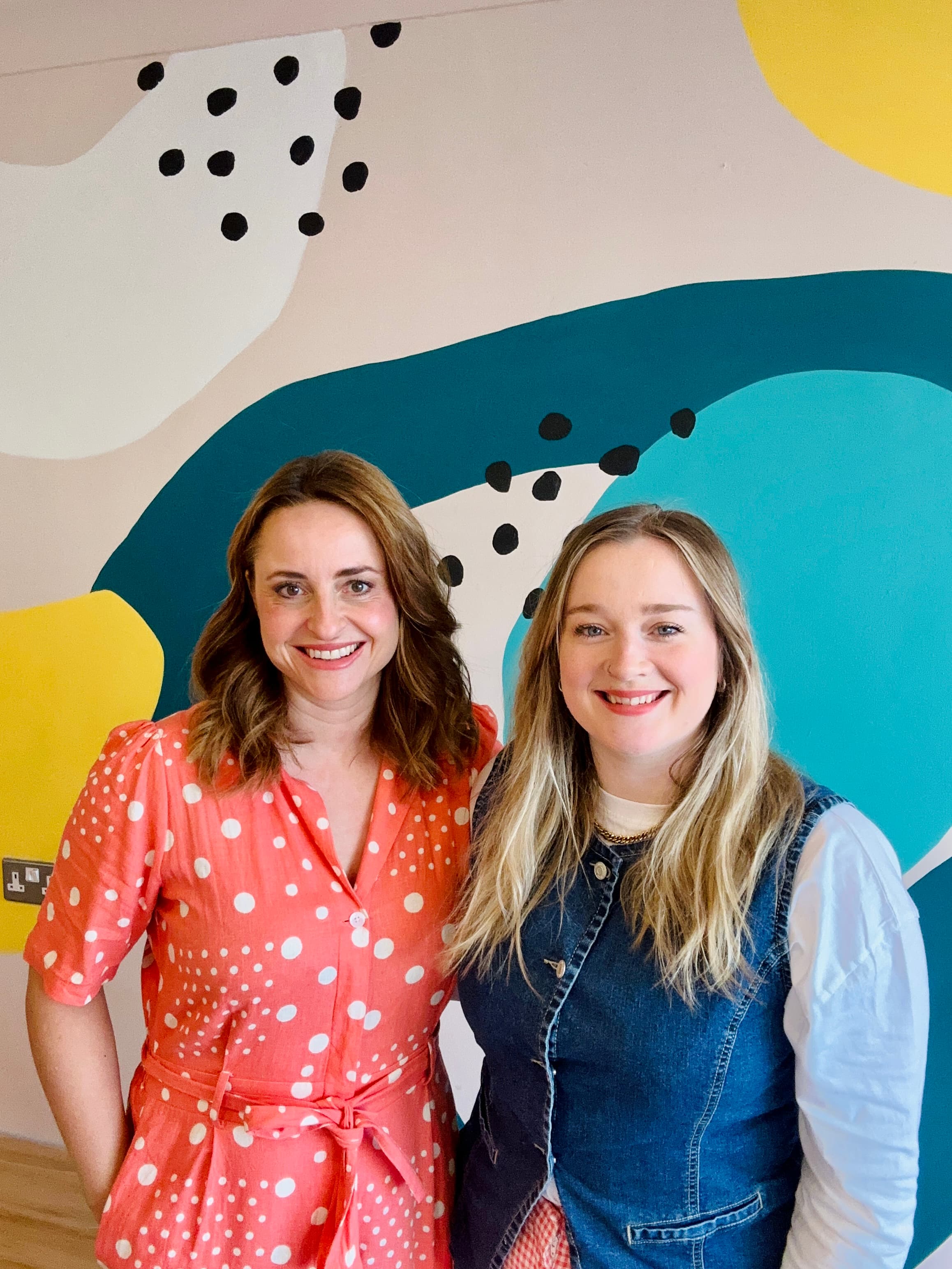 Two women stand smiling in front of a colorful mural featuring abstract shapes and polka dots, in a bright café setting.